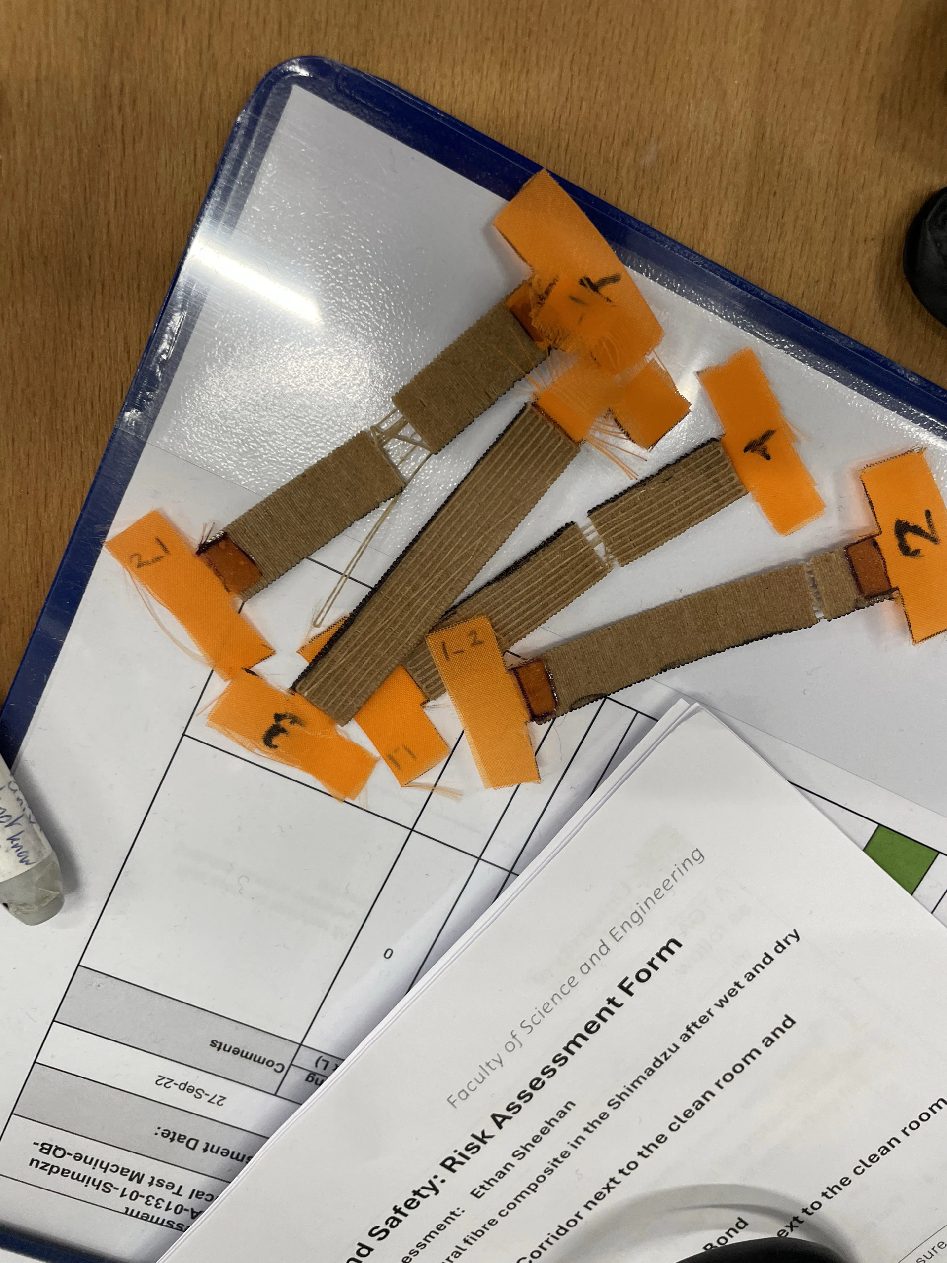 Five tape-labelled flax/PLA strips on a tray, some frayed at the ends, lying on the risk assessment form for the Shimadzu tests