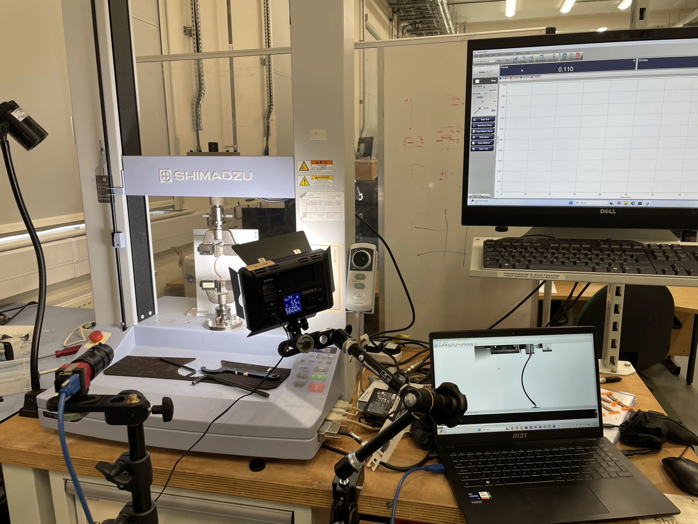 Closer view of the Shimadzu machine with the LED light panel, curvature-tracking camera and a laptop showing the specimen outline