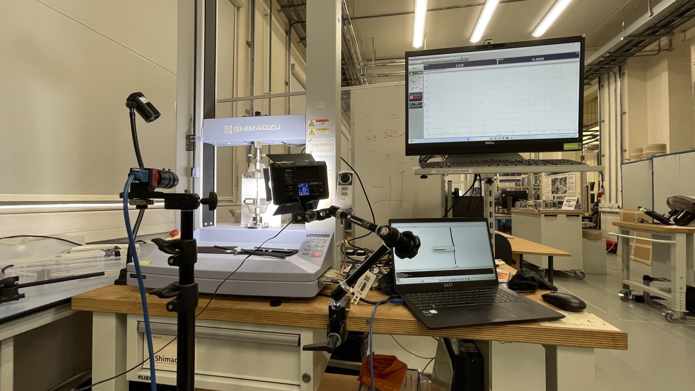 Shimadzu tensile machine on a bench with the camera arm, LED panel, acquisition laptop and a live force-displacement trace on the monitor