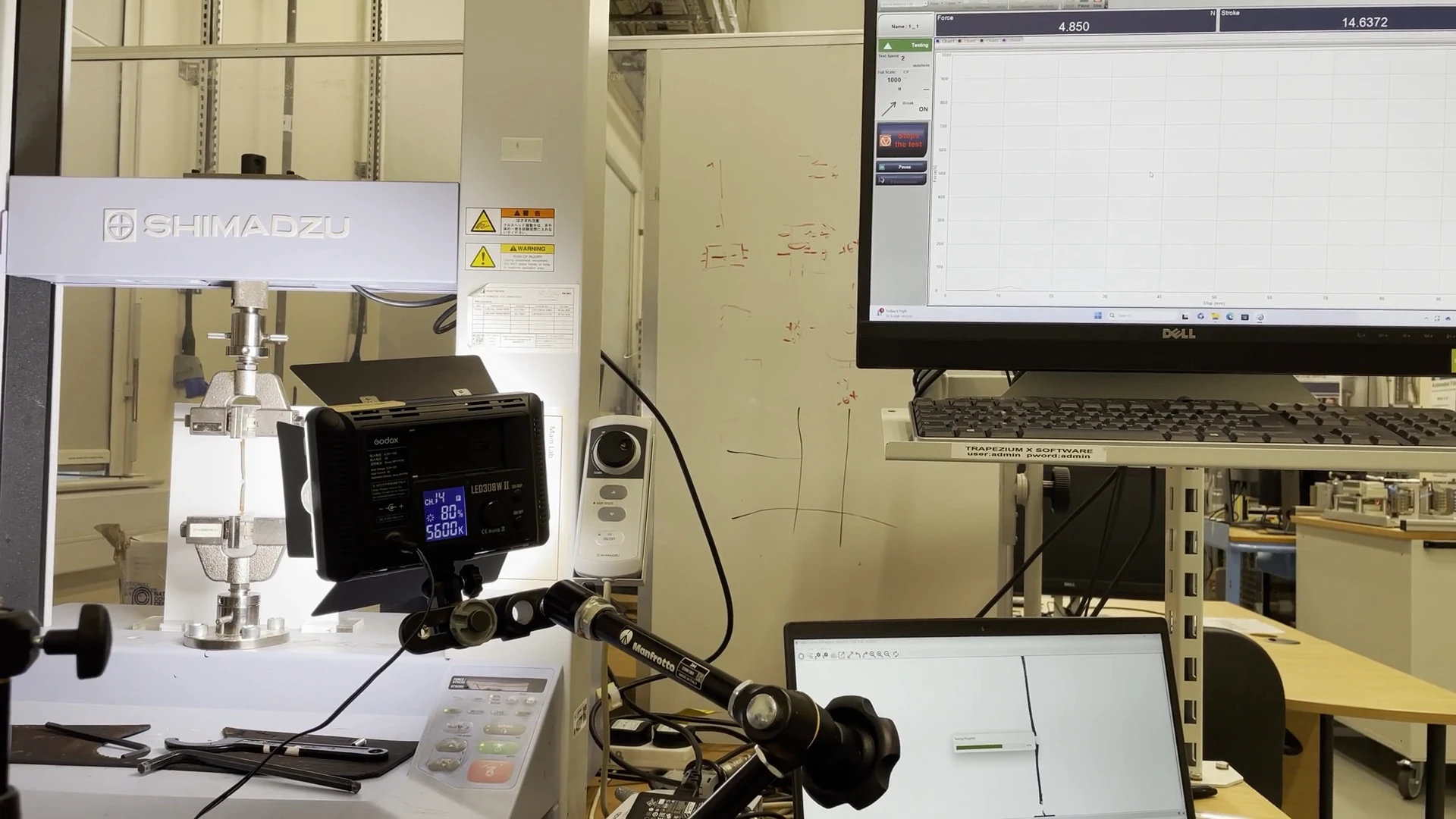 Pan across the tensile rig during a live test, from the Shimadzu frame to the acquisition monitor