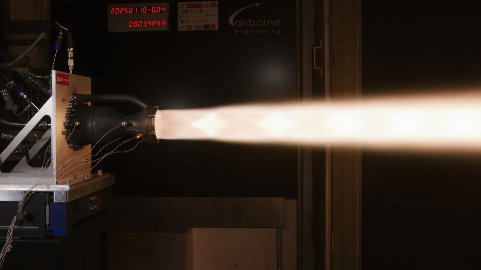 Engine 1 at steady state on the AEL thrust stand, a bright plume with Mach diamonds streaming right across a dark test cell