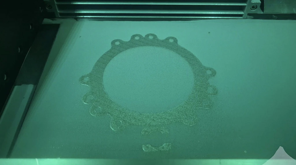 Build-camera frame of the powder bed mid-print with the fused cross-section of the chamber flange standing out against fresh grey powder