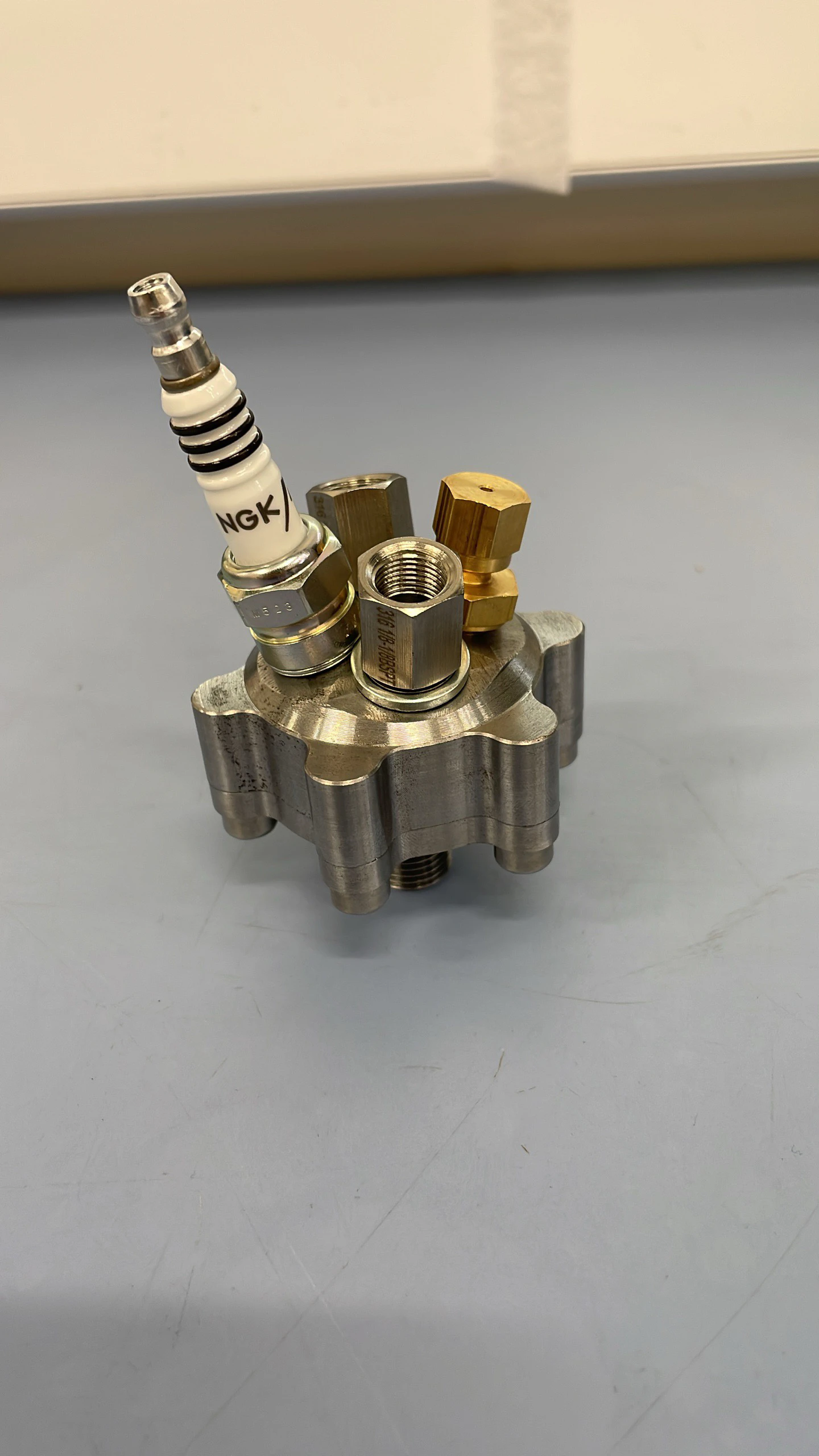The assembled igniter standing on a bench, spark plug pointing up from the steel body with two brass inlet fittings.