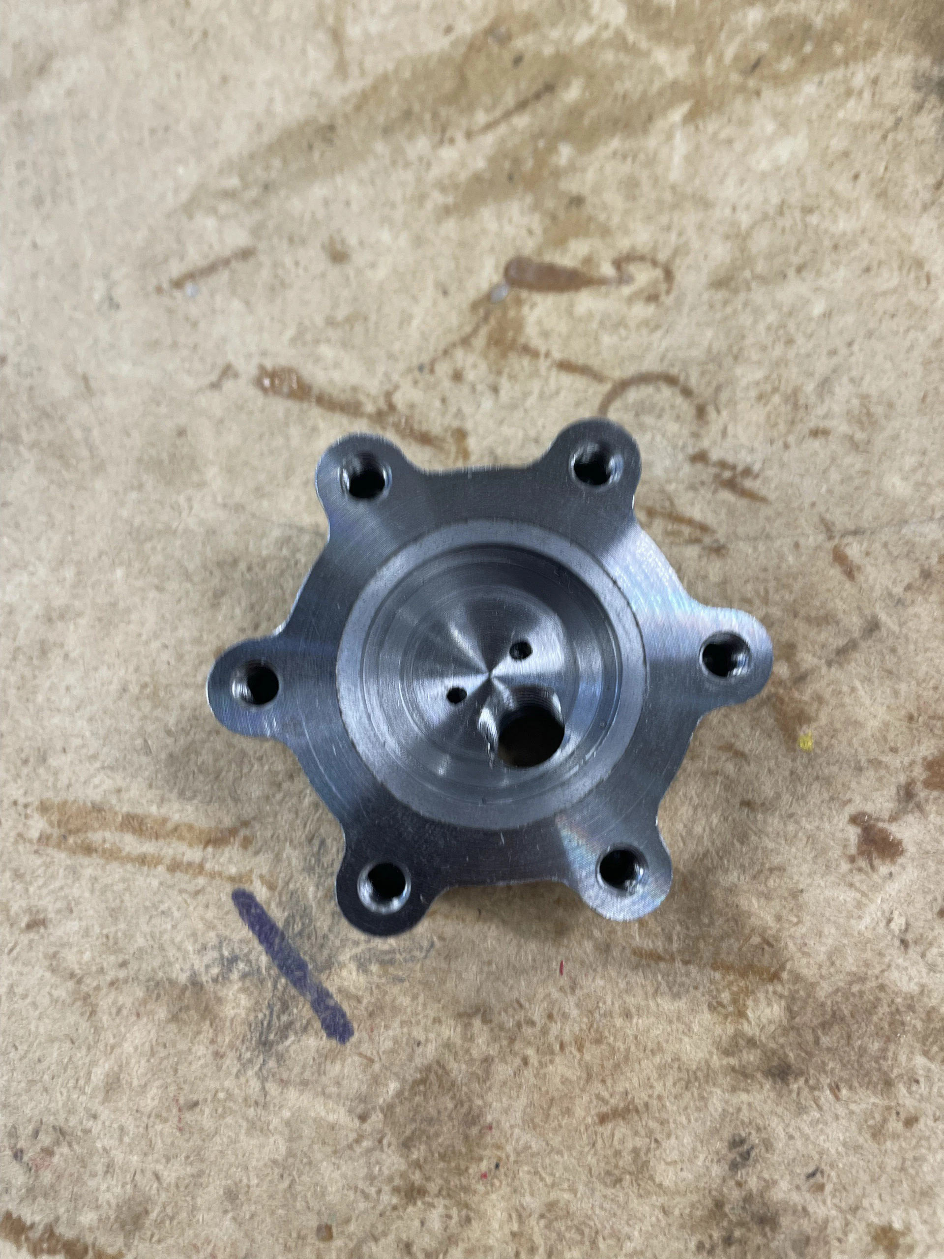 Hexagonal steel flange with six bolt holes and a central port lying on a workshop bench.