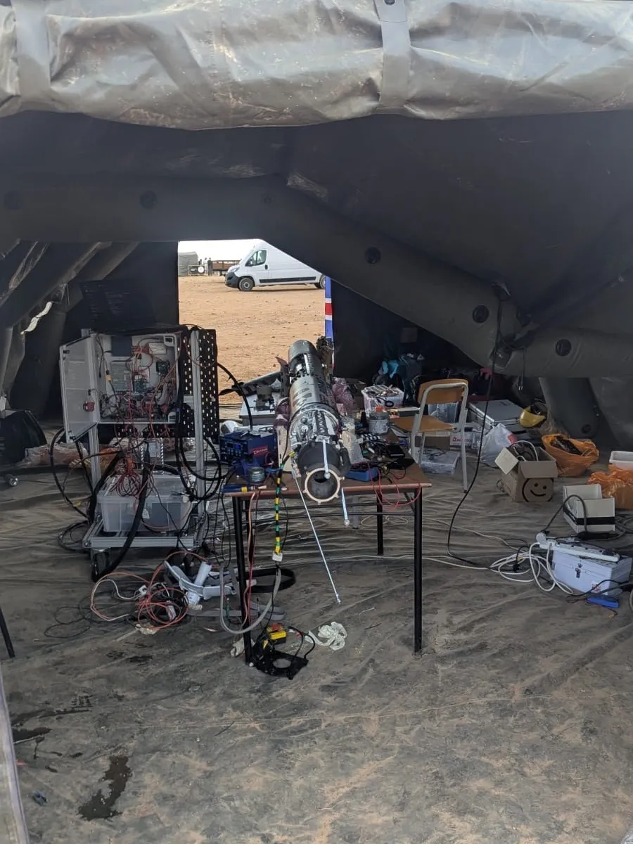 The engine assembly on a trestle table in the launch site tent, feed lines running to the ground support cart behind it, a van and open red earth visible through the tent doorway