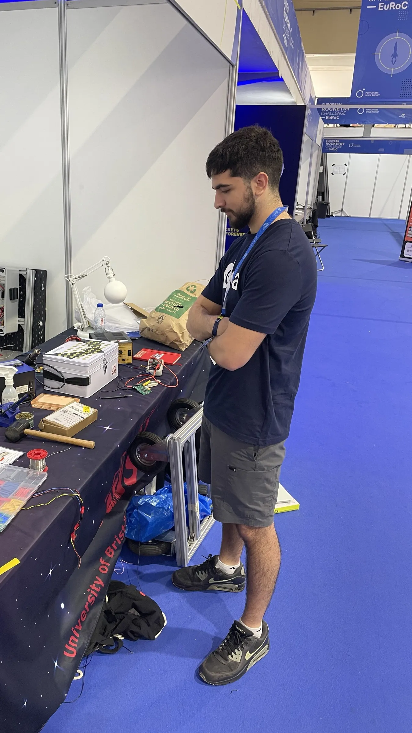 The team pit inside the EuRoC hangar, a team member at a work bench of wiring, connectors and tools with an avionics enclosure open behind it, the competition aisle and banners beyond