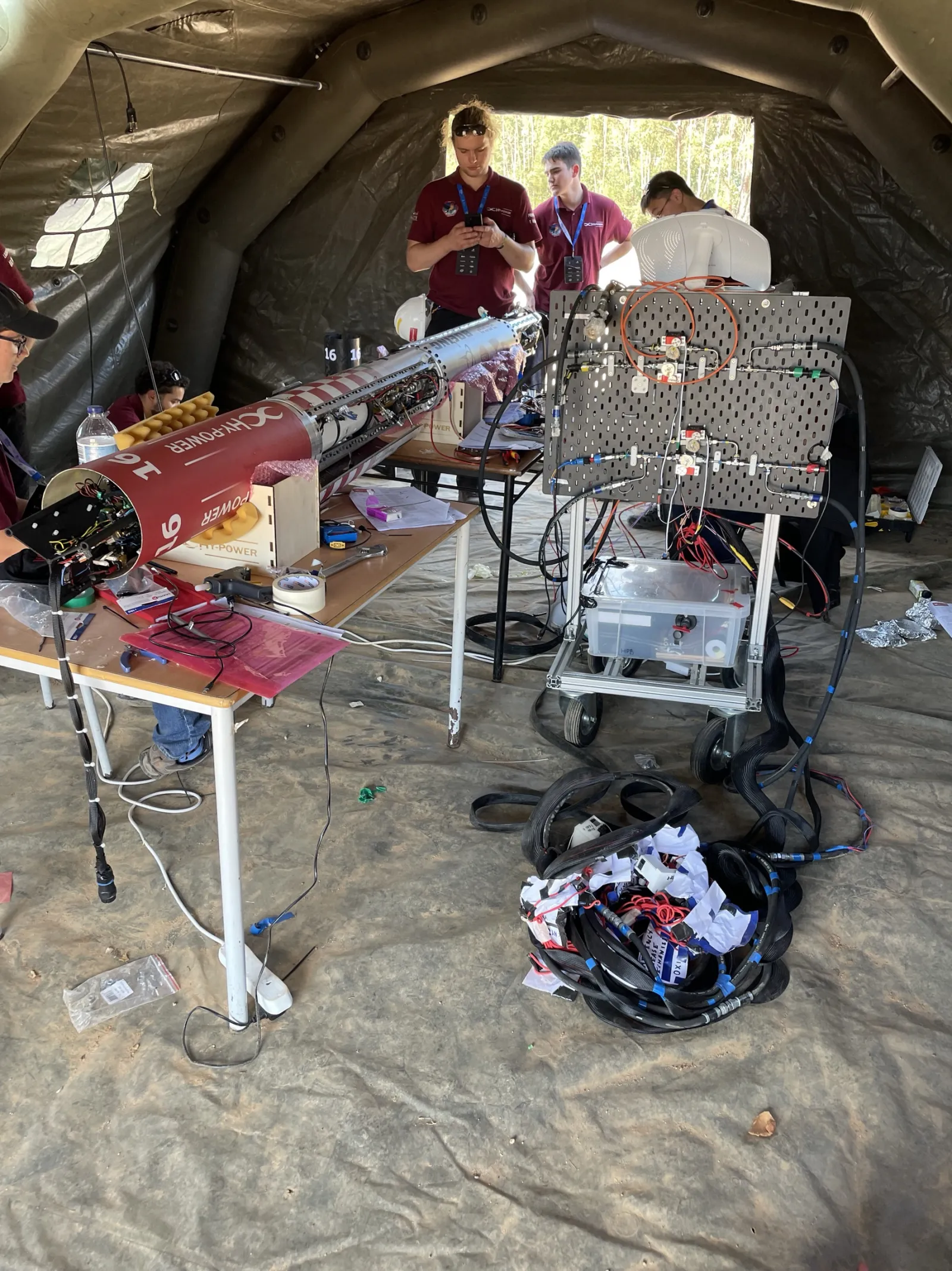 Inside an inflatable tent at the launch site: Waxwing open on trestles with its harness exposed, the ground support panel of valves and fittings on a wheeled frame beside it, umbilical hoses coiled on the dust floor and team members working around it