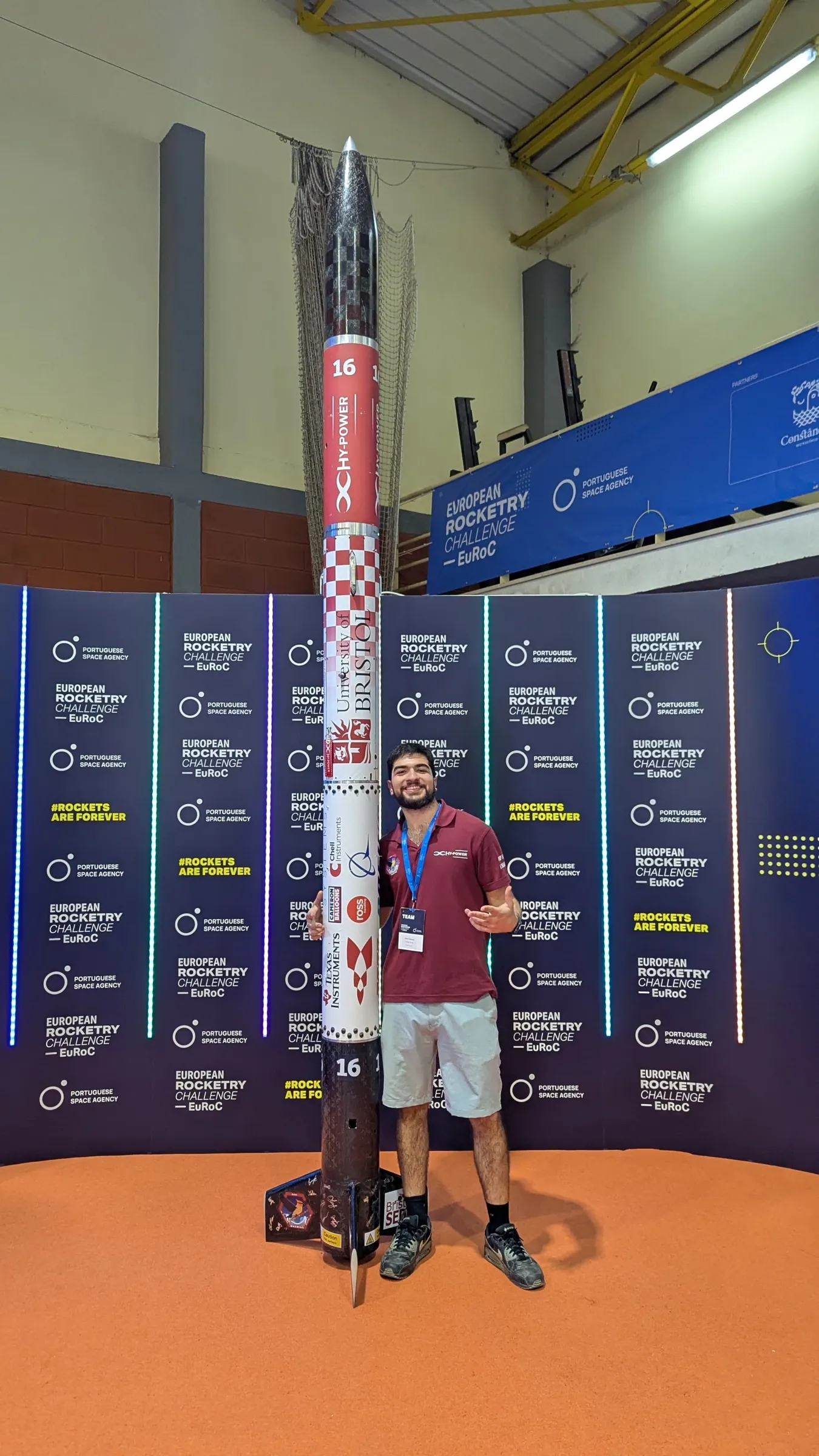 A HyPower team member in a maroon team polo standing beside Project Waxwing, held vertical against the EuRoC step and repeat backdrop inside the competition hangar
