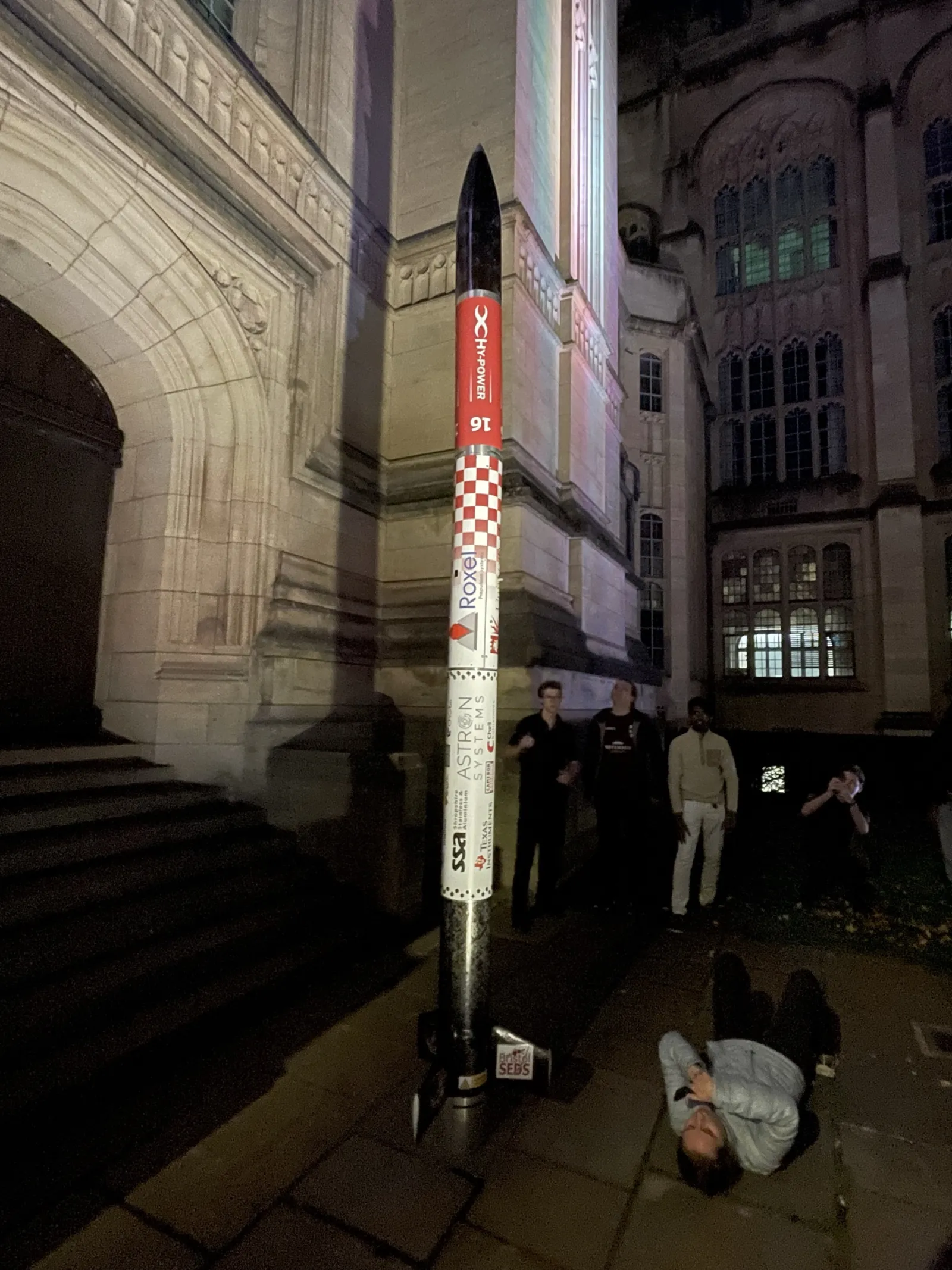 Project Waxwing standing vertical at night outside the Wills Memorial Building in Bristol, sponsor decals down the airframe, team members alongside and one lying on the paving looking up at the nose