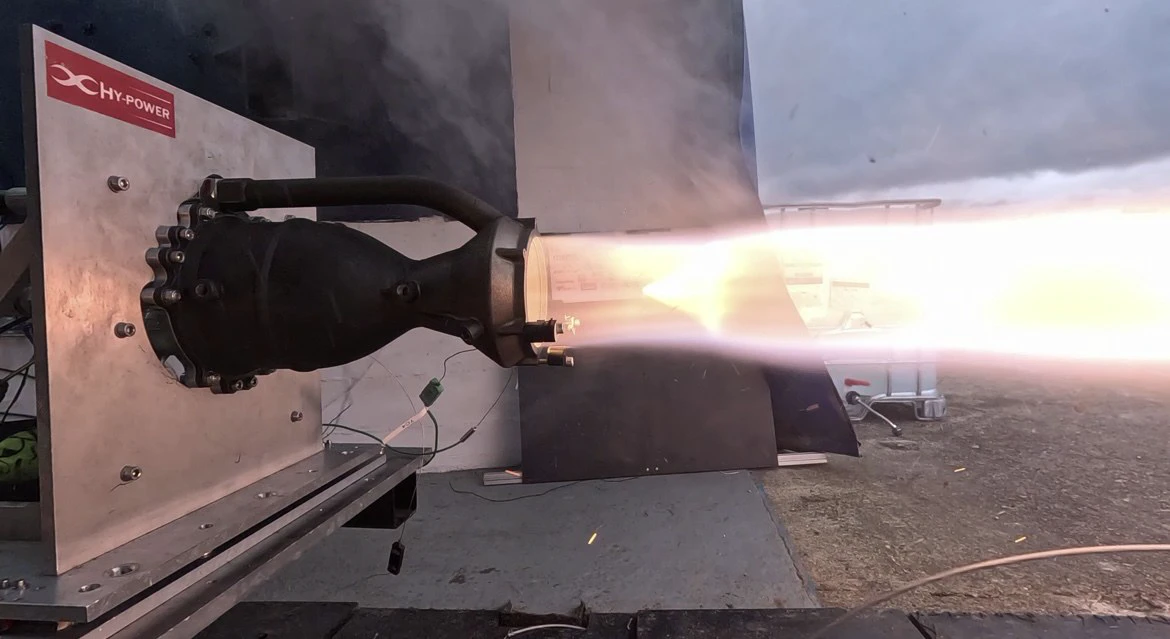 Engine 1 bolted horizontally to the Astron test stand under a HyPower placard, plume streaming right during a burn