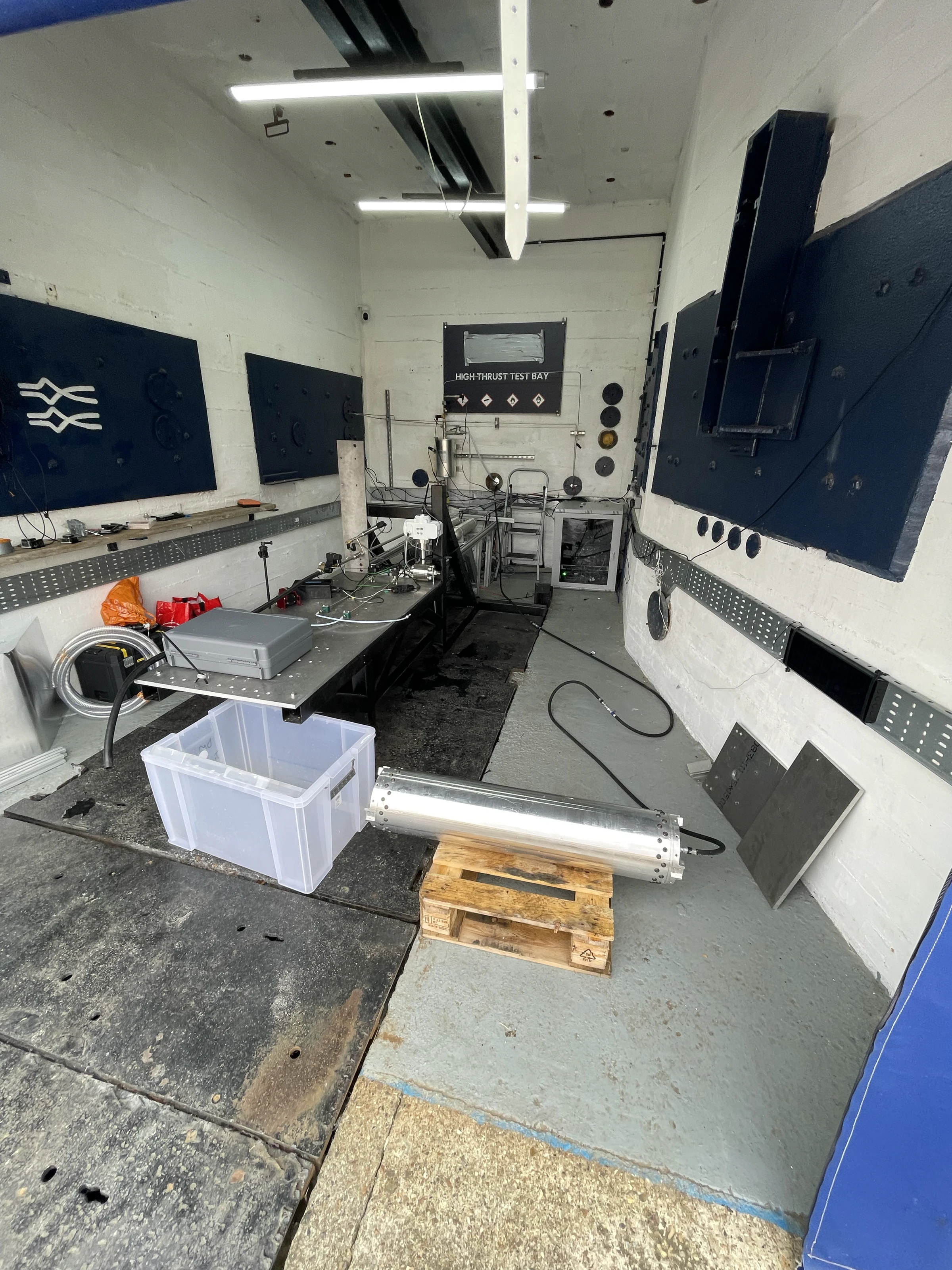 The empty high-thrust test bay at Astron: white walls, dark acoustic panels, a bare stand and the tank lying in a crate on the floor