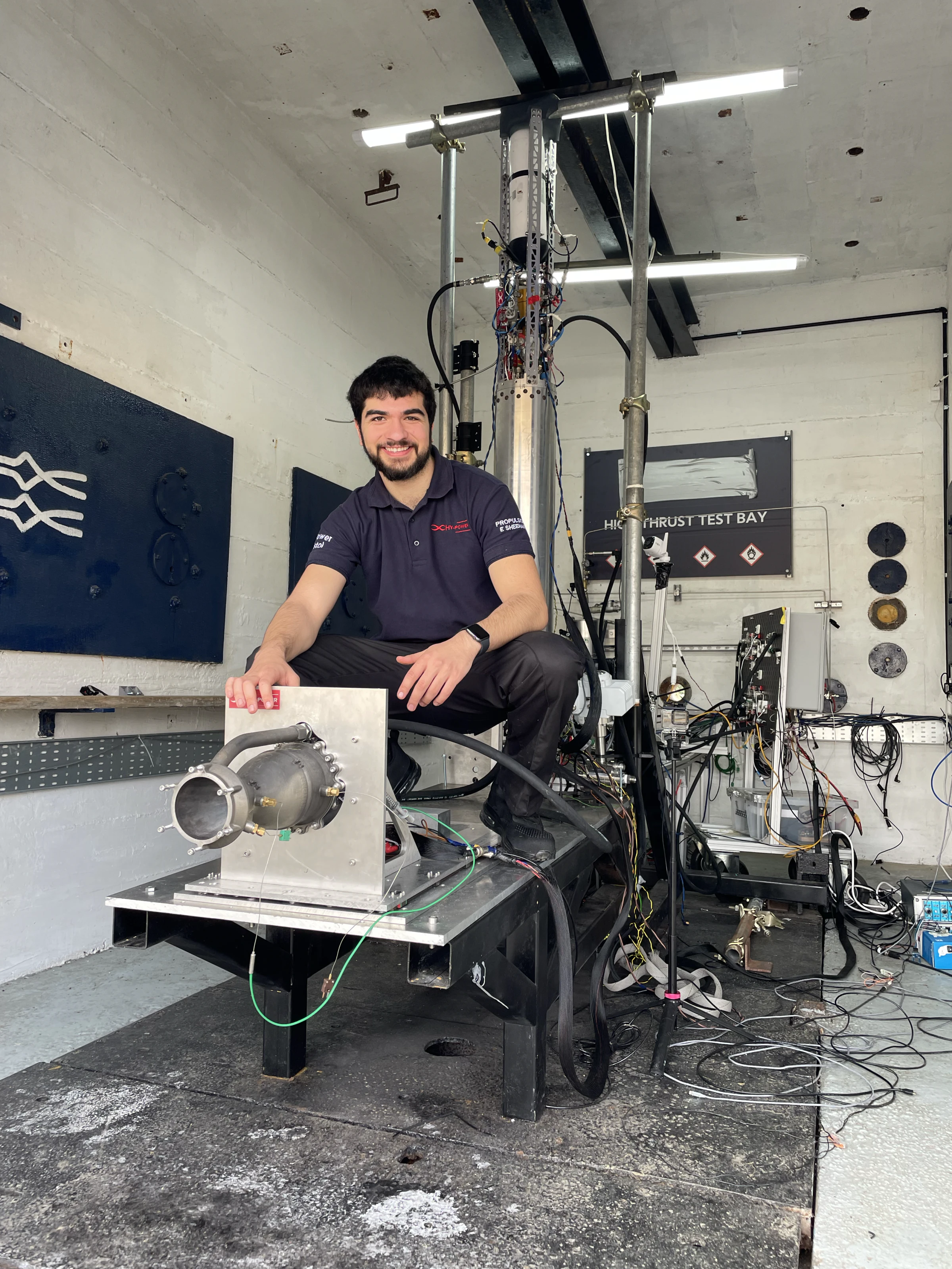Ethan Sheehan sitting beside Engine 1 on its mounting plate in the test bay, the vertical tank and its plumbing behind him