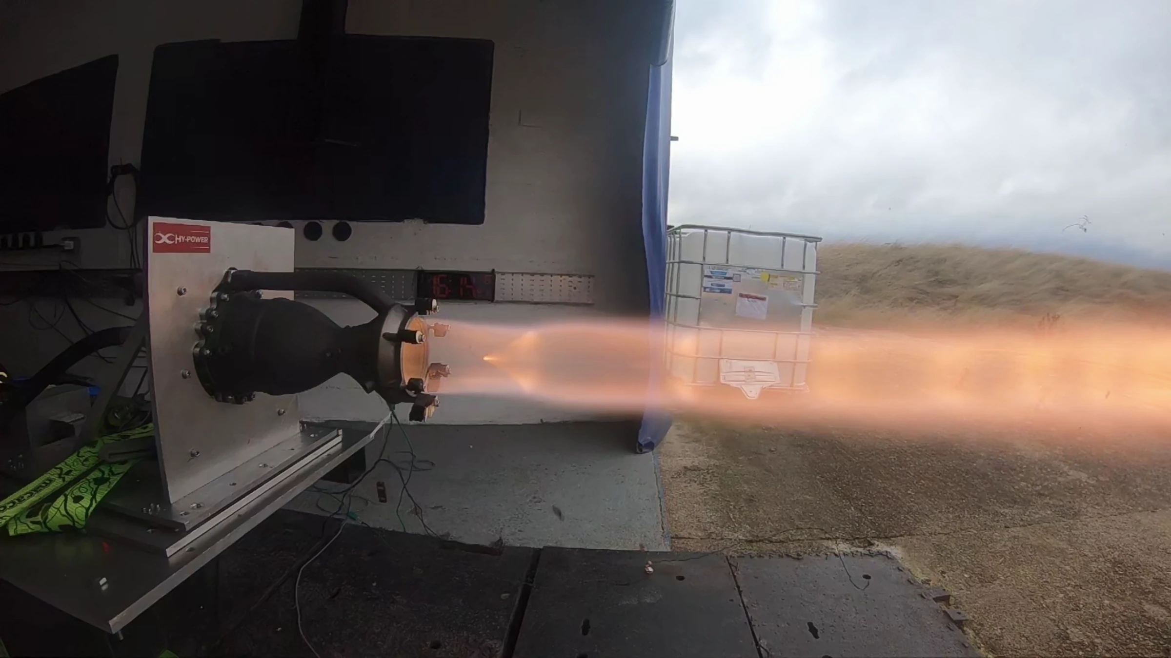 Hotfire 1 running: a translucent orange plume streaming right from the nozzle under an overcast sky