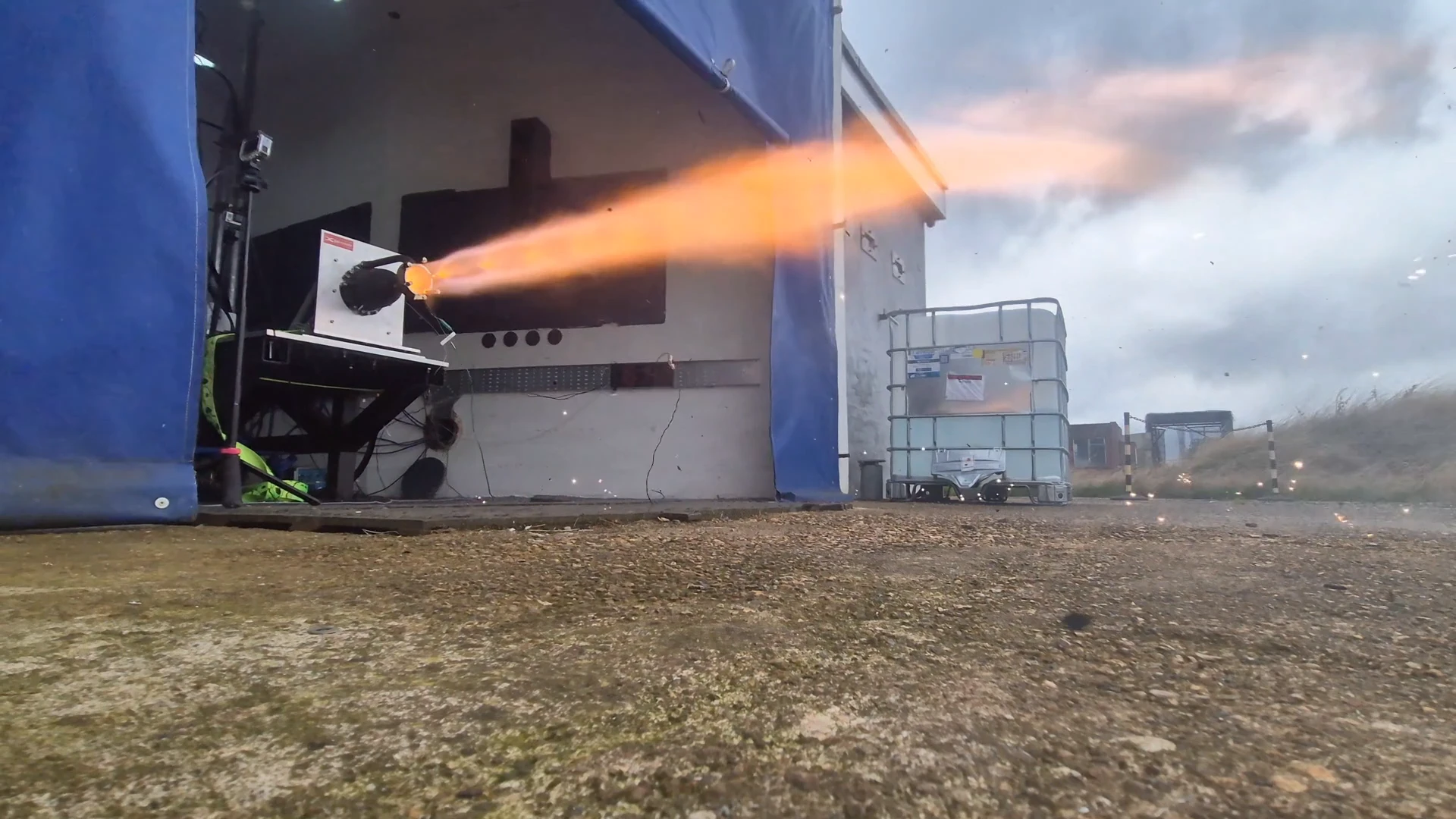 Wide 4K view of the test bay from outside: the sparkler ignites in a shower of sparks, a short orange plume streams out of the bay, then white vapour as the oxidiser runs out