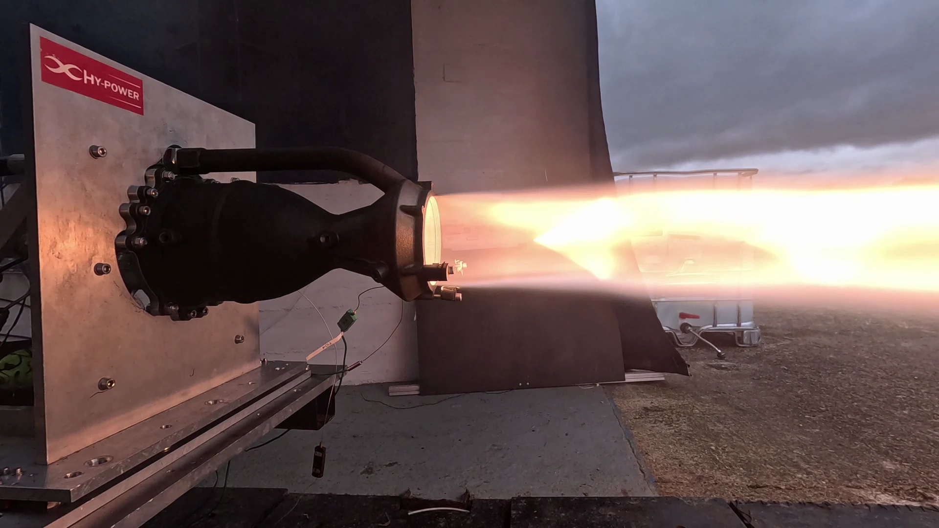 Engine 1 firing horizontally on the Astron test stand at dusk, a long golden plume with a bright shock core against a grey evening sky