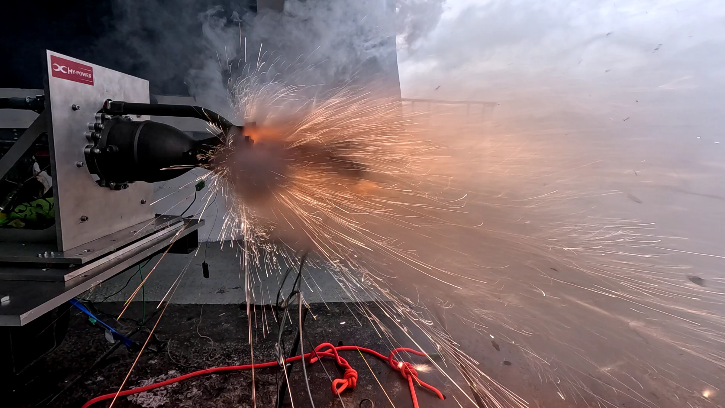 Hotfire 3 ignition: a burst of sparks at the nozzle against a dark test pad, red hose coiled in the foreground