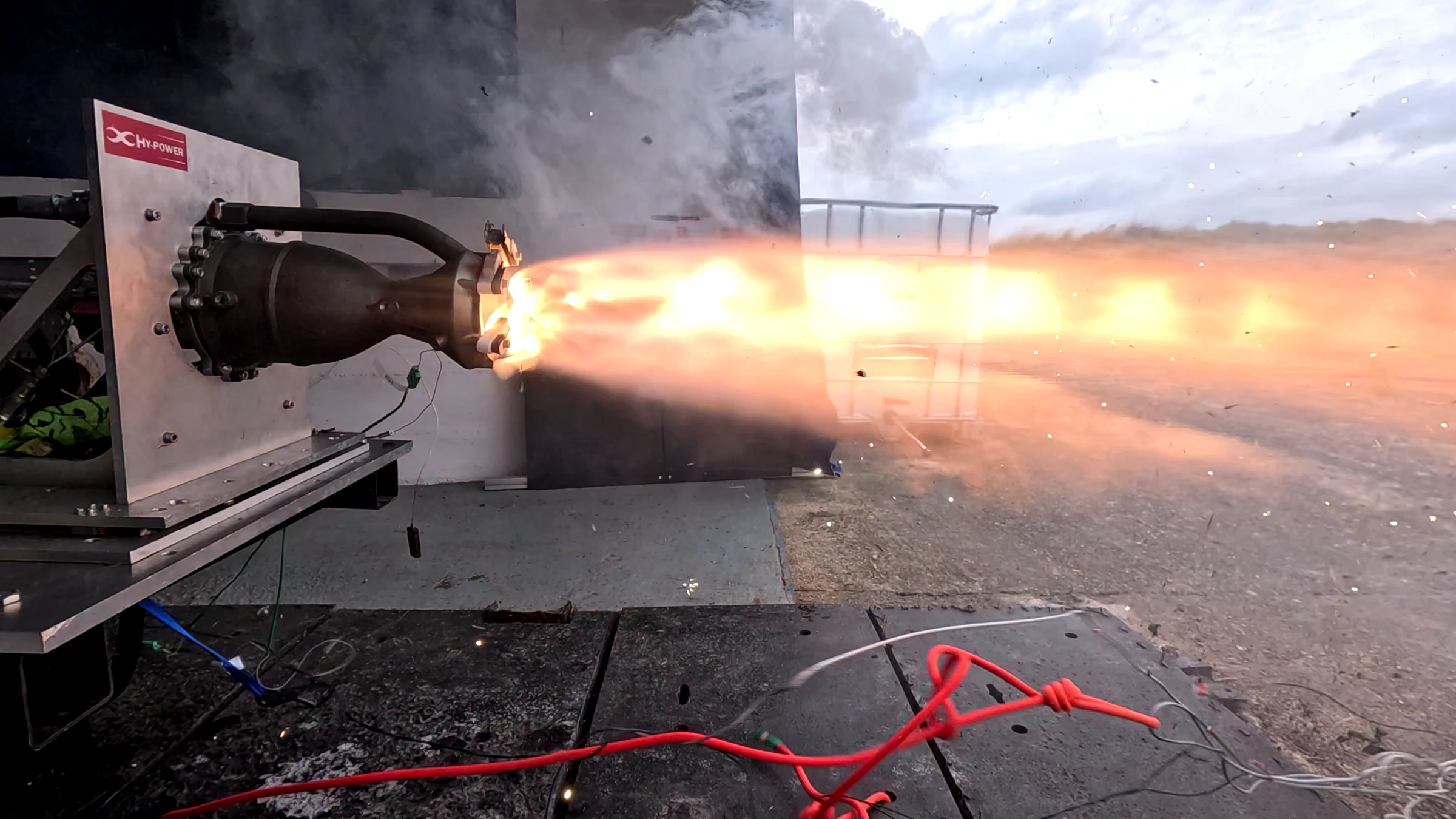 Hotfire 3 at full chamber pressure: a dense plume with a clear shock train leaving the nozzle, the ground dark and the sky pale behind it