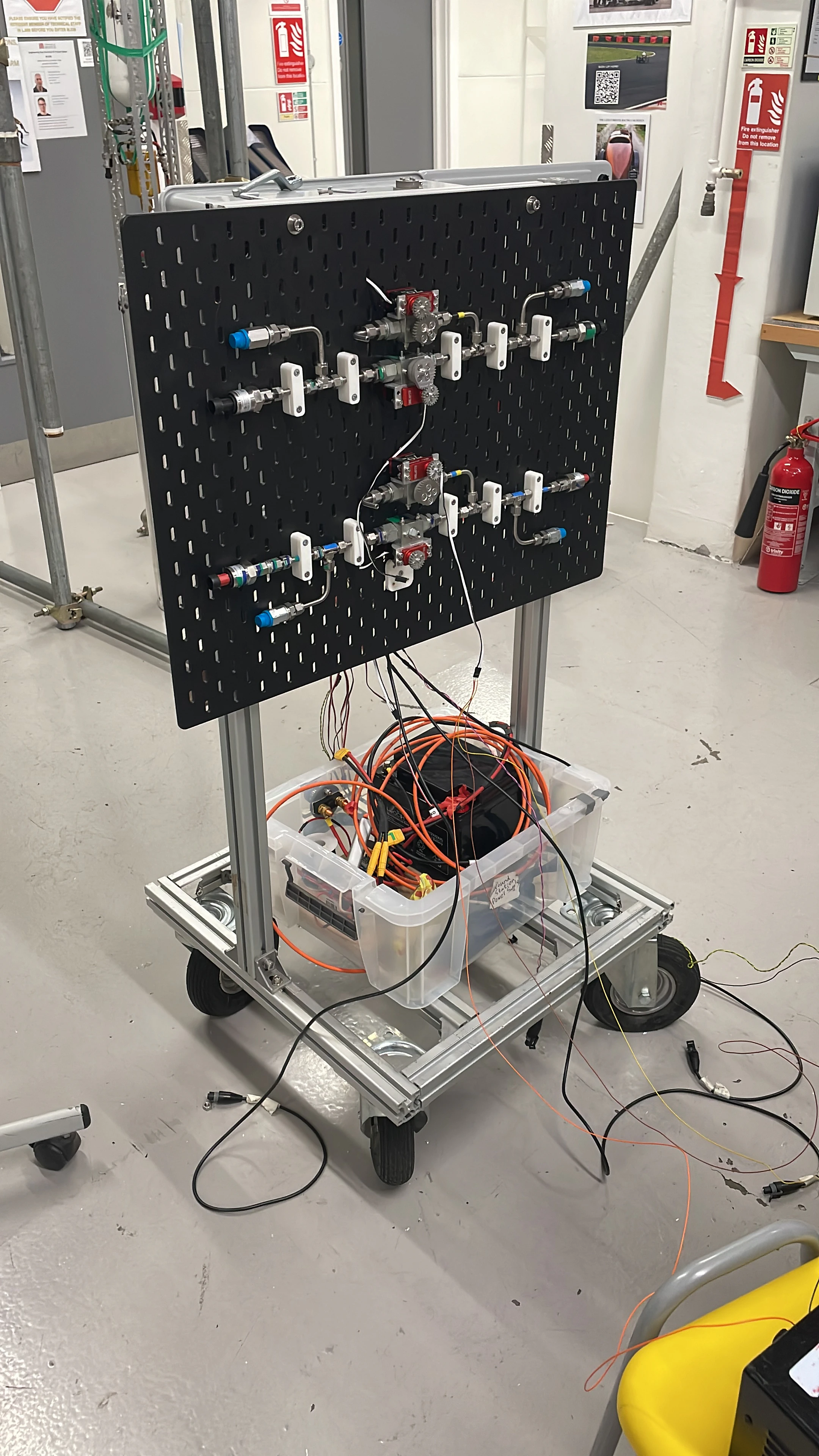 The fluid control panel on a trolley: a black perforated board carrying two rows of hand valves and solenoids, with a control box in a clear case underneath