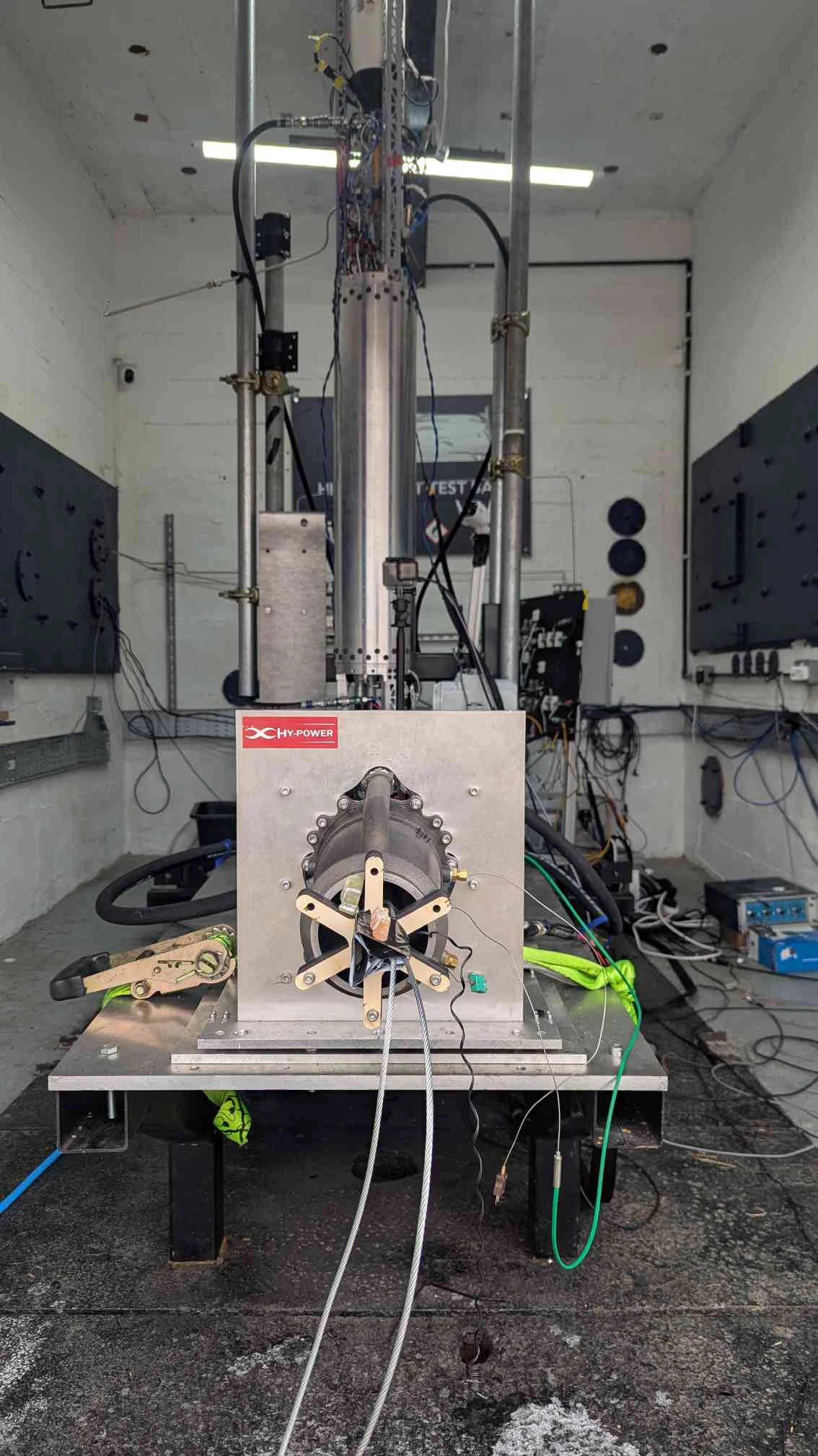 The 2025 stack in the Astron test bay: the flight tank standing vertical against the back wall, Engine 1 bolted horizontally to a plate at floor level behind a HyPower placard