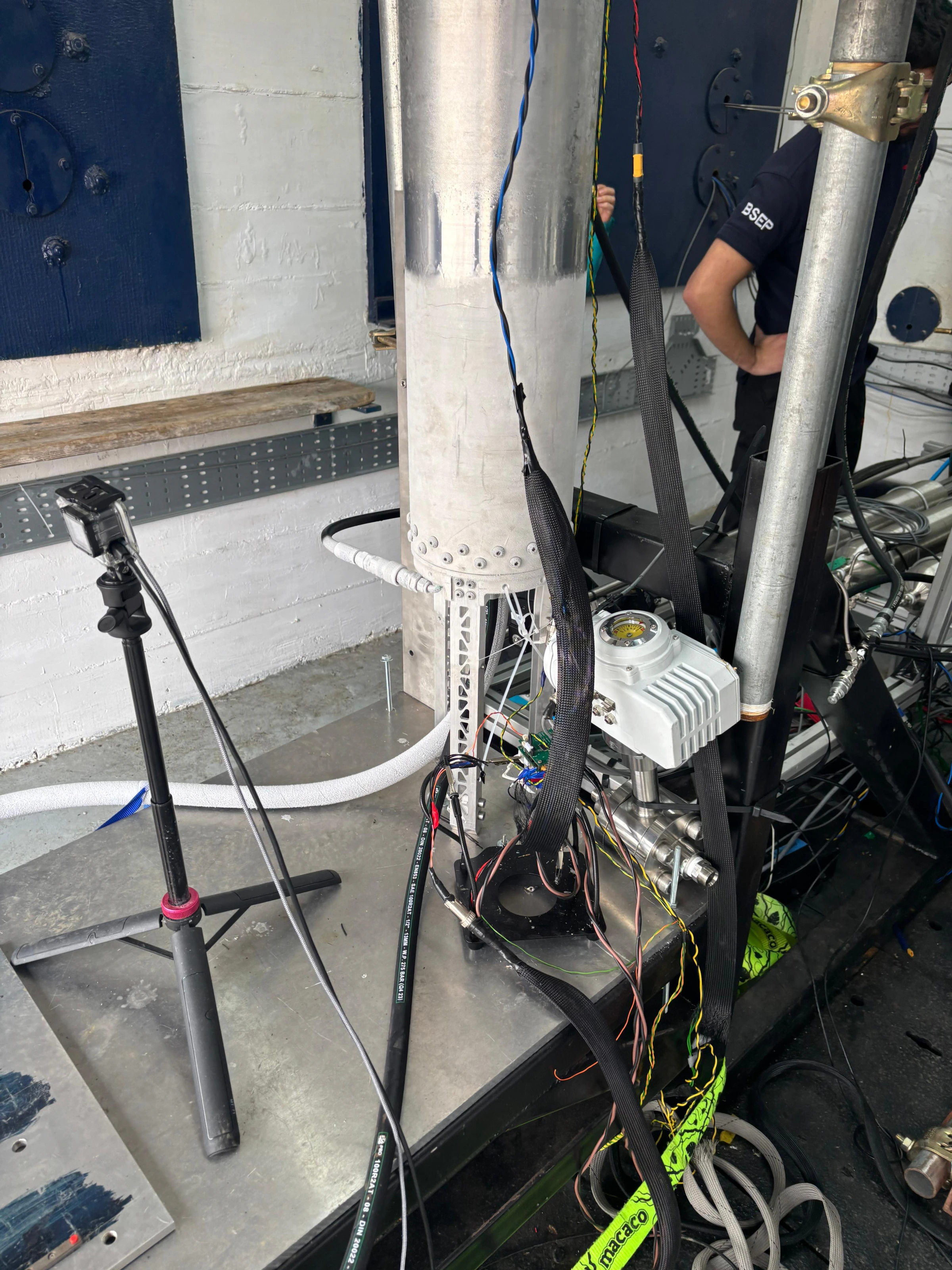Close on the base of the vertical flight tank, wrapped in instrumentation cable with a camera tripod and pressure lines around it