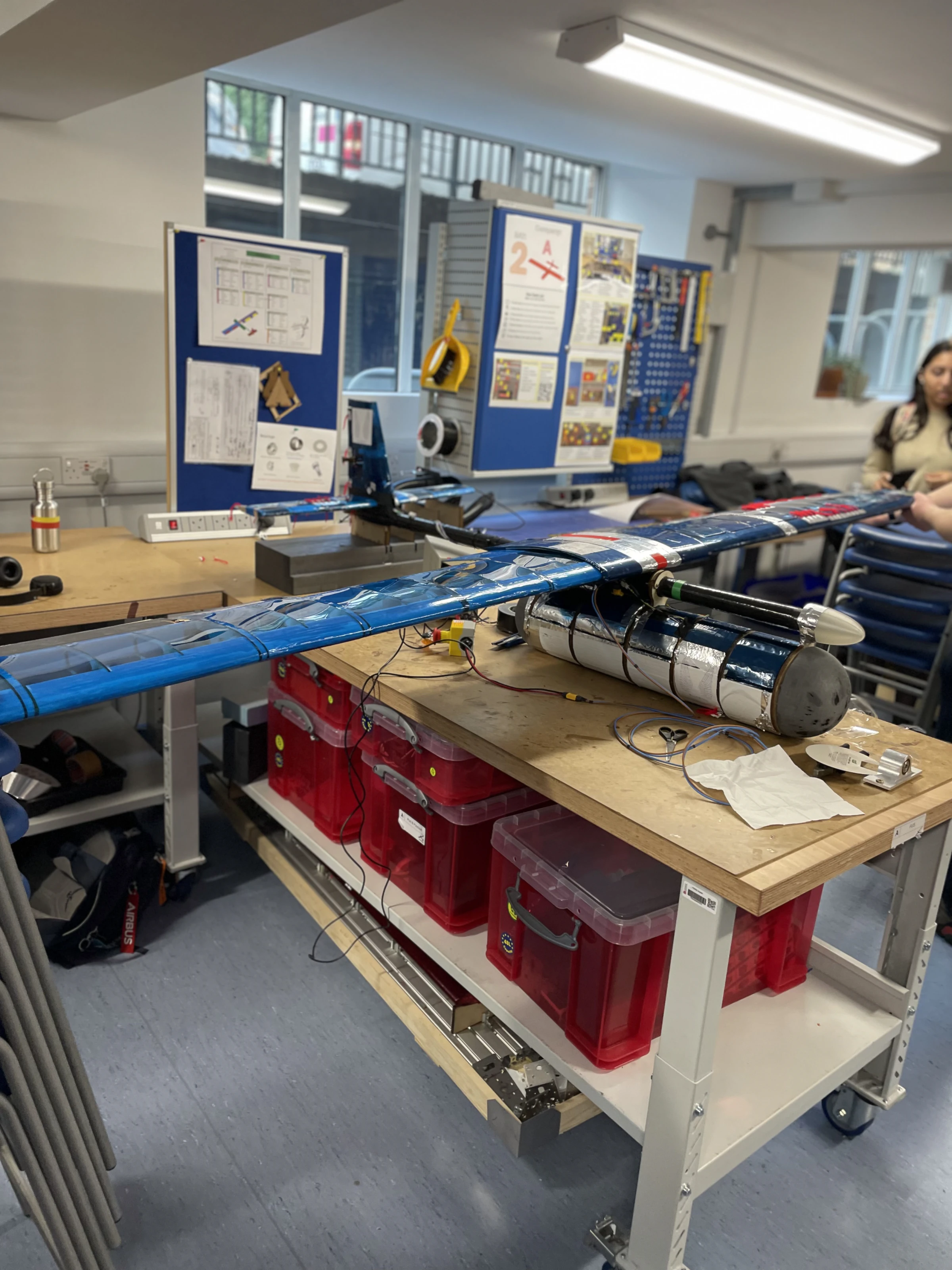 The complete UAV on a bench trolley: full-span blue wing, silver instrument pod slung under the carbon fuselage spar, tail at the far end