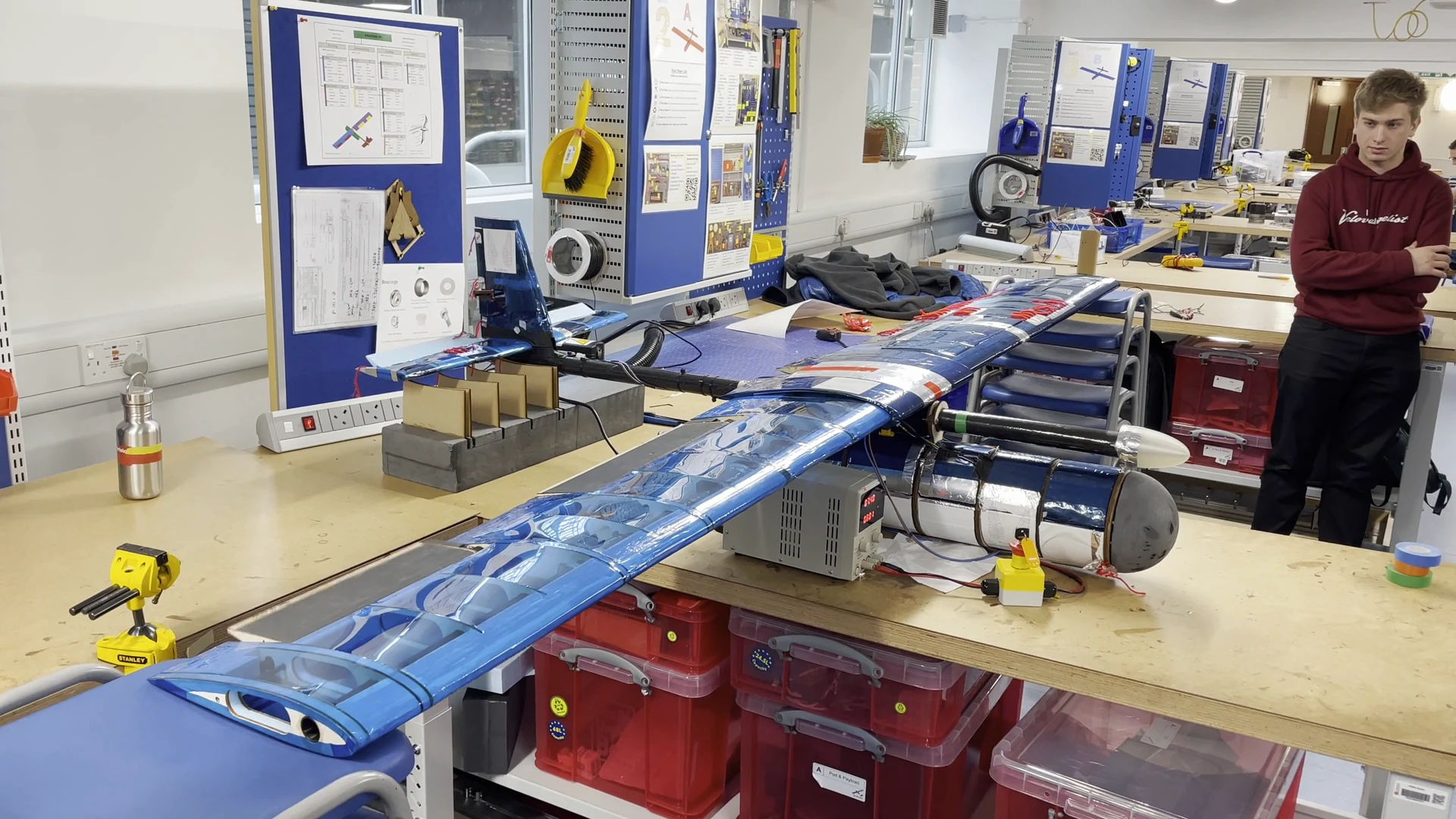 The finished BlueBird UAV on a bench trolley in the lab: full blue film-covered wing across two benches, silver instrument pod under the carbon fuselage spar, students in maroon lab coats working around it