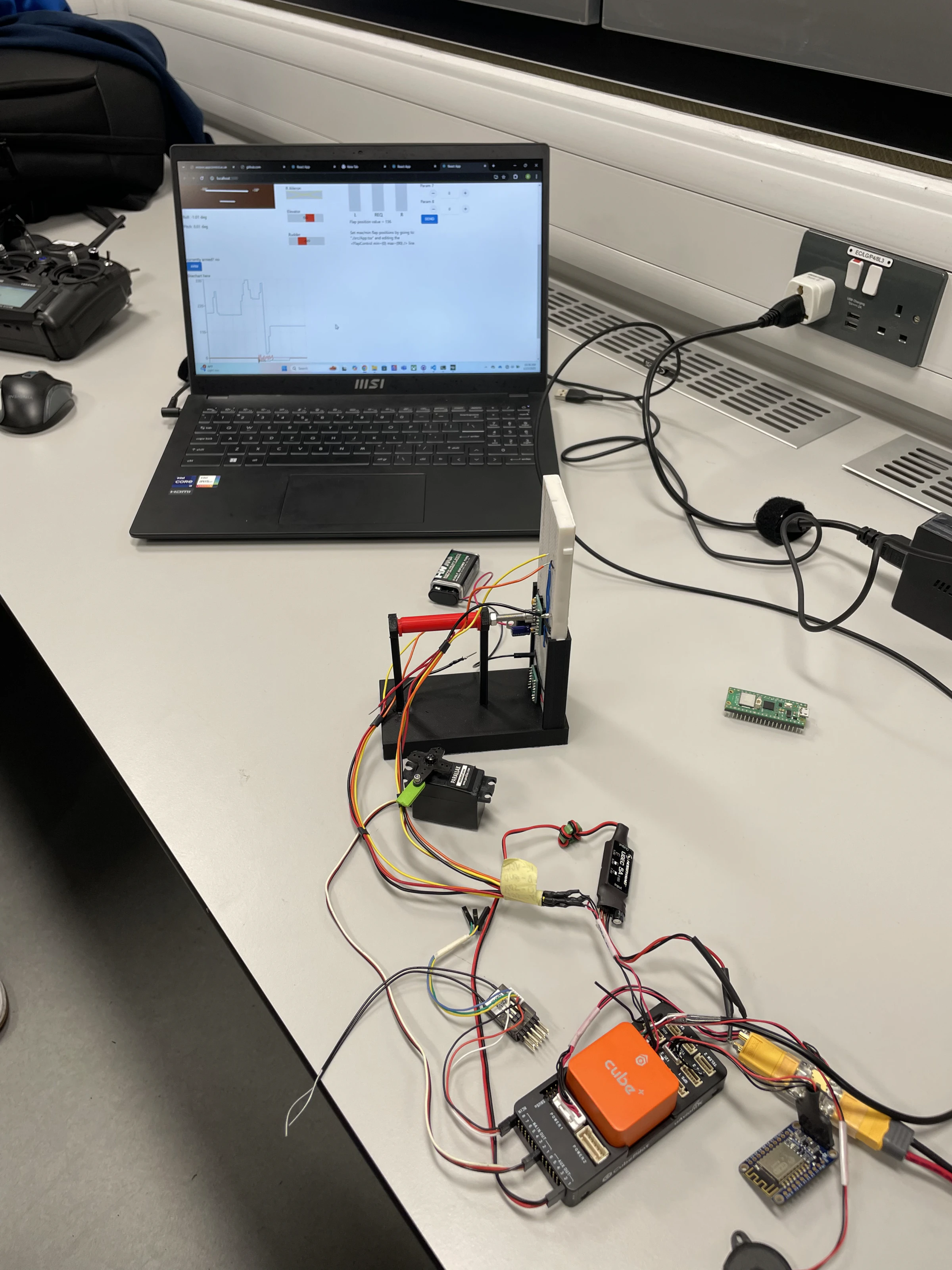A bench with a laptop running the ground station, a servo on a black test stand, a Cube Orange flight controller, an ESP8266 Wi-Fi module and a radio transmitter, all wired together