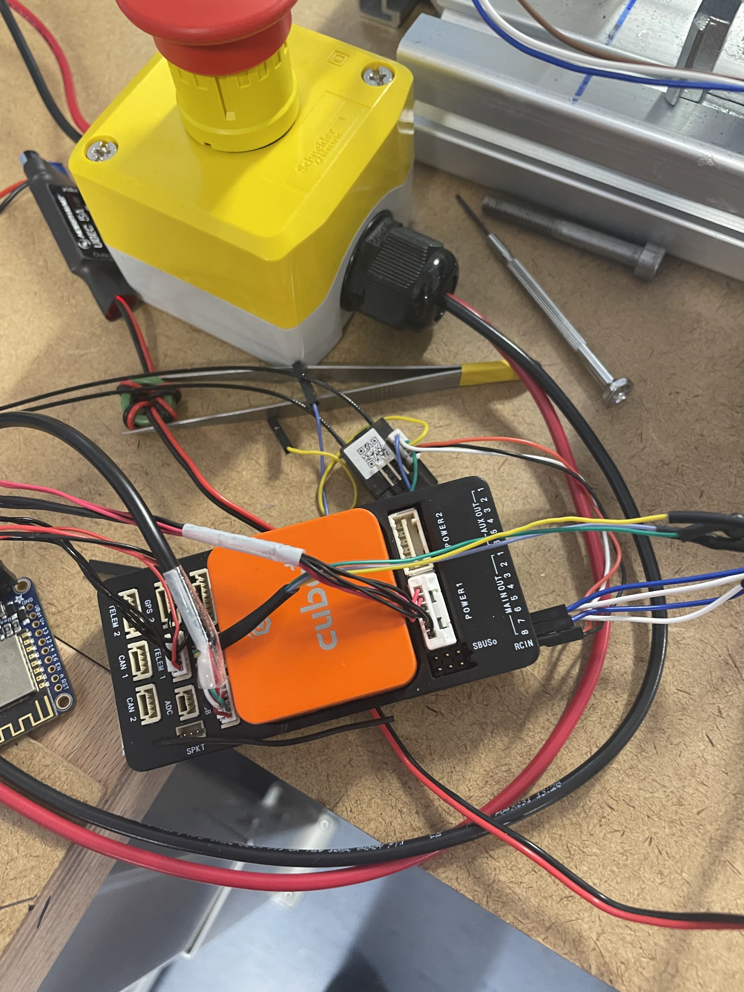A Cube Orange flight controller in its orange case with telemetry and Wi-Fi modules, power wiring and a yellow emergency-stop box on the bench