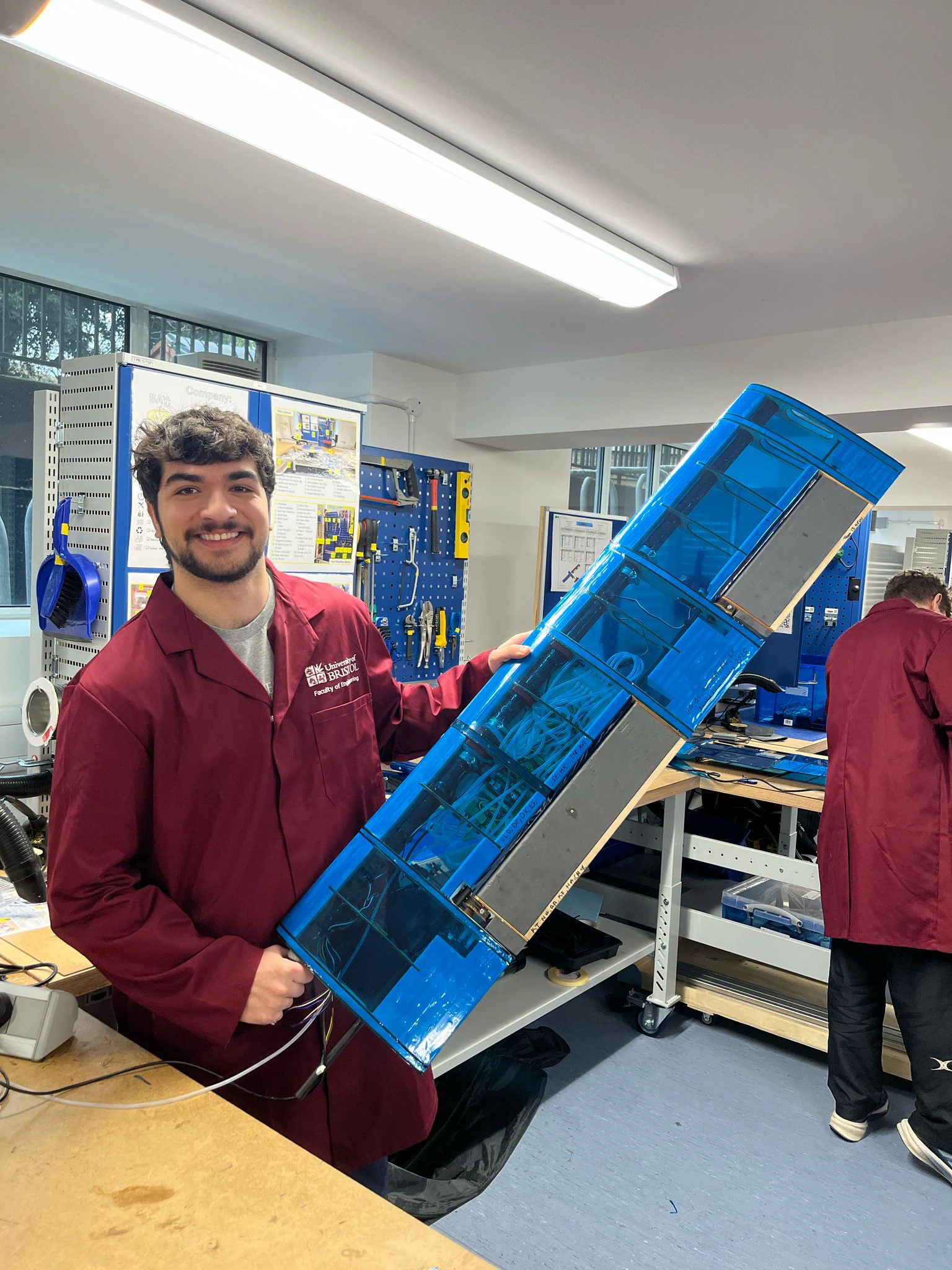 Ethan in a maroon Bristol lab coat holding the blue film-covered wing upright in the workshop, grinning