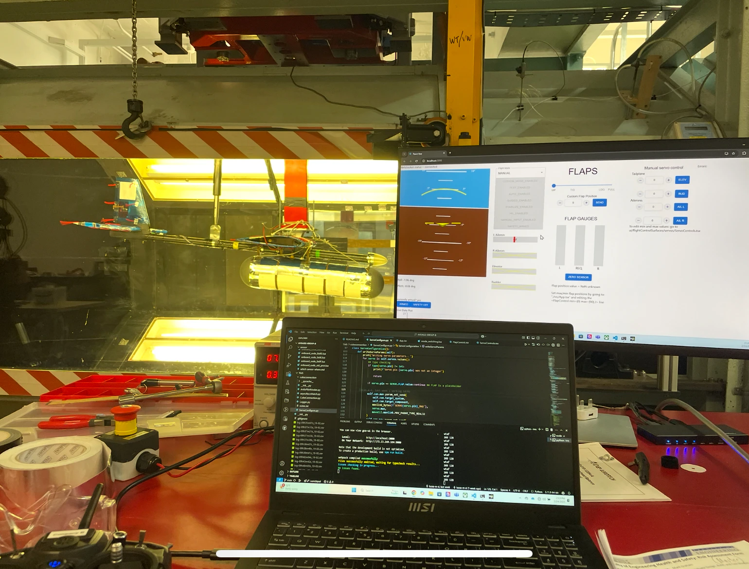 The tunnel test section with the fuselage inside, a monitor showing the ground station FLAPS page and a laptop showing the telemetry code running in an editor