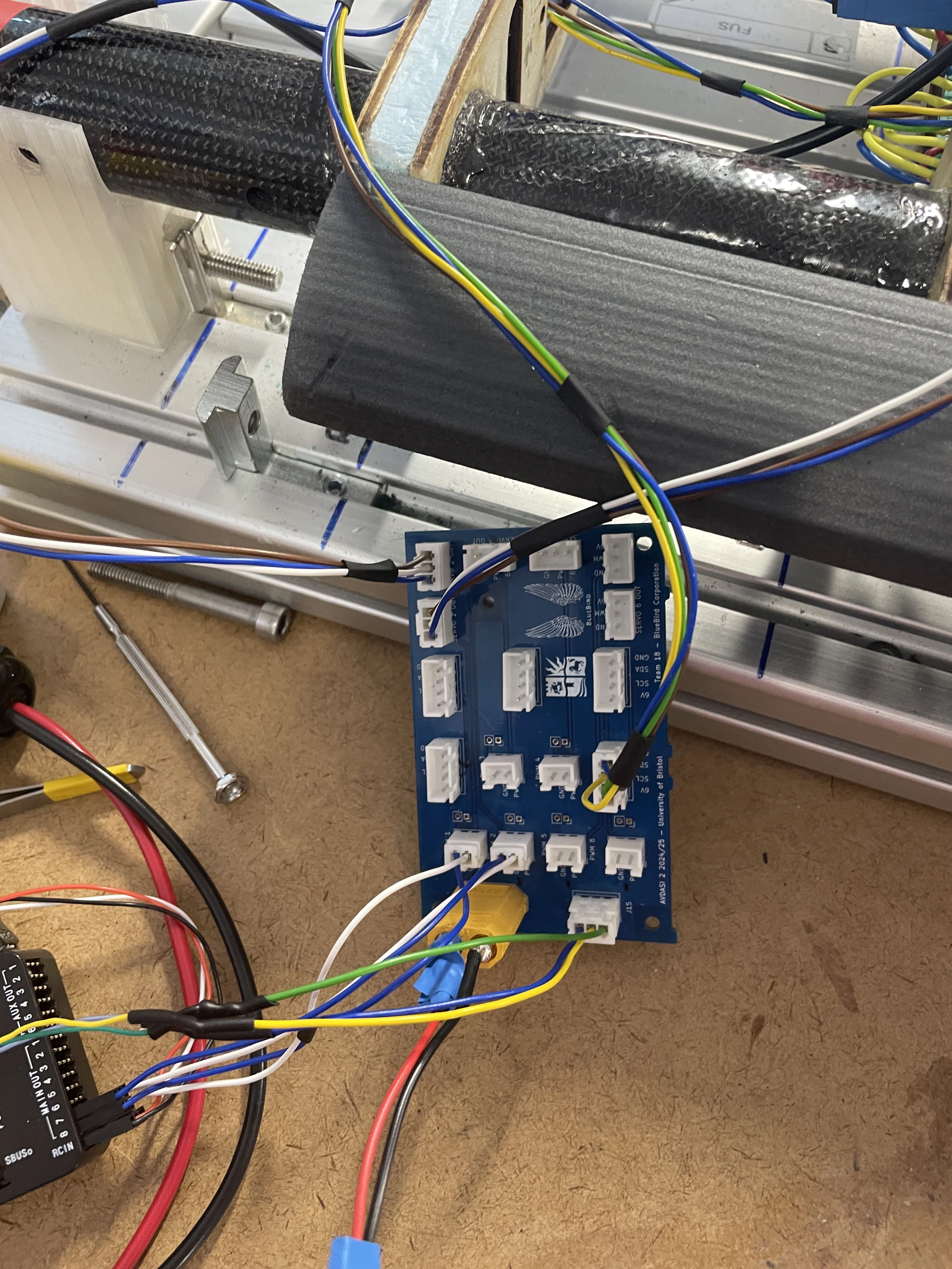 The blue harness distributor circuit board on the workbench beside the carbon fuselage spar, wired with heavy red, black, yellow and green cables