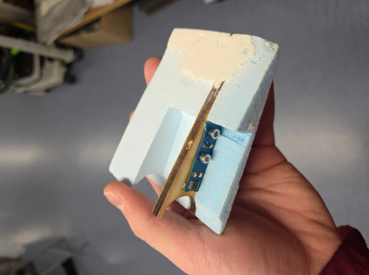 A hand holding a foam wing-section mock-up with the angle sensor board and its brass arm bonded in place