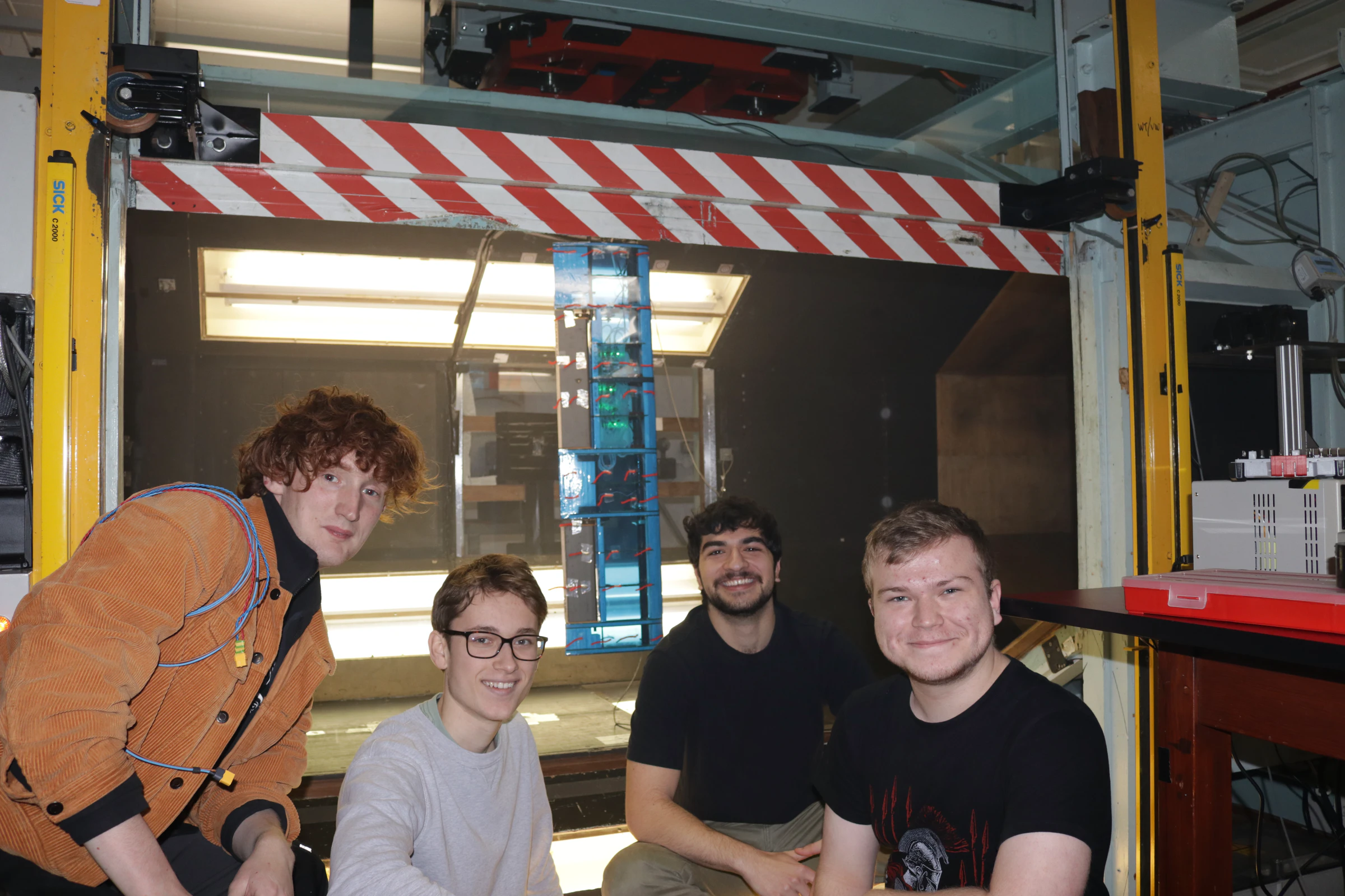 Four members of Team 18 crouched in front of the open wind tunnel test section, the tufted blue wing standing vertically inside it with the green sensor LEDs lit