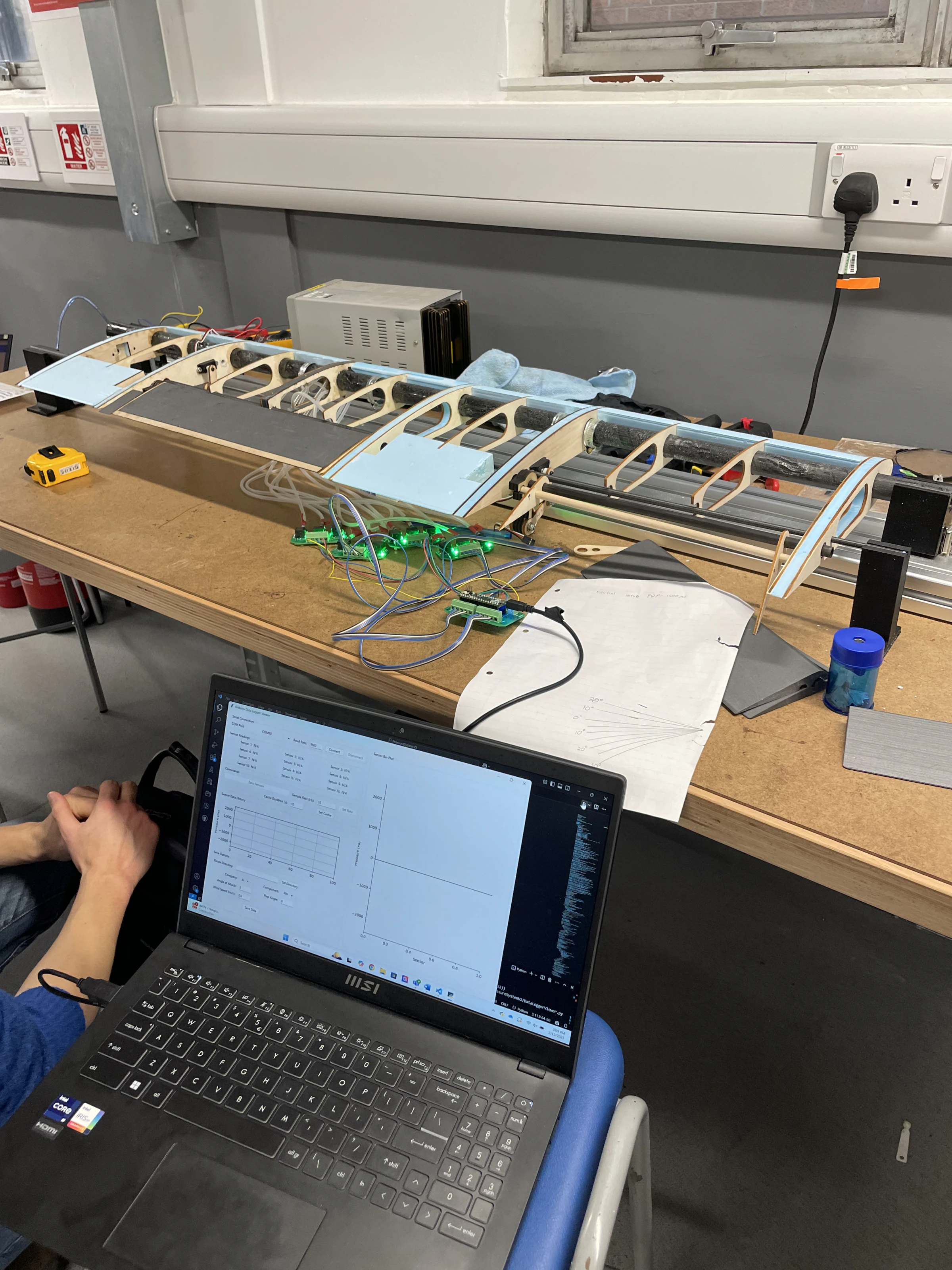 Wing structure open on the workshop bench with green sensor boards mounted at the flap hinges and a laptop showing a telemetry plot