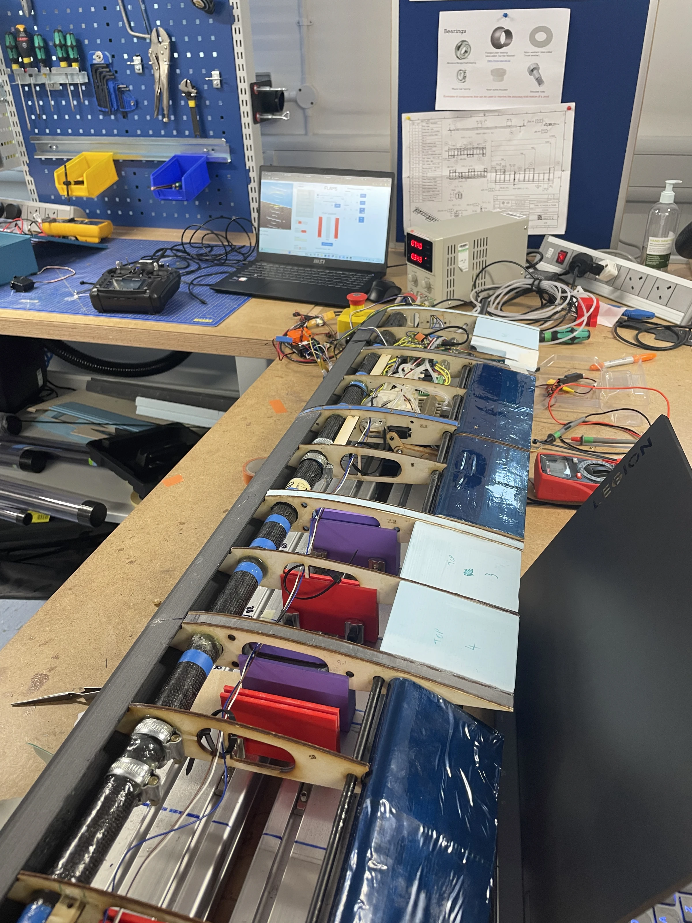 A long workshop bench holding the wing sections with the pressure acquisition system, coloured manifolds and a laptop, blue pegboard behind